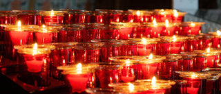 Rows of red glass candle holders with lit candles, glowing with warm orange and yellow flames against a dark background.