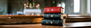 A stack of Liturgy of the Hours books with red, black, and dark blue spines sits on a wooden table in a blurred indoor setting with warm bokeh lights in the background.